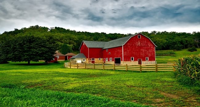 A vibrant red barn with weathered wooden fence bordering a lush green field under a dramatic cloudy sky, evoking rustic homestead charm.