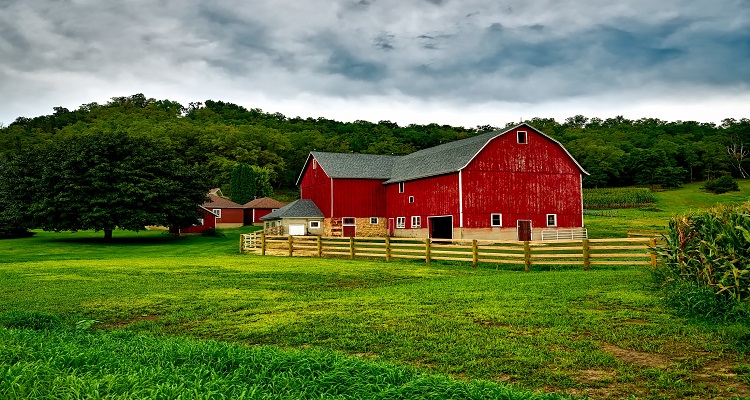 A vibrant red barn with weathered wooden fence bordering a lush green field under a dramatic cloudy sky, evoking rustic homestead charm.